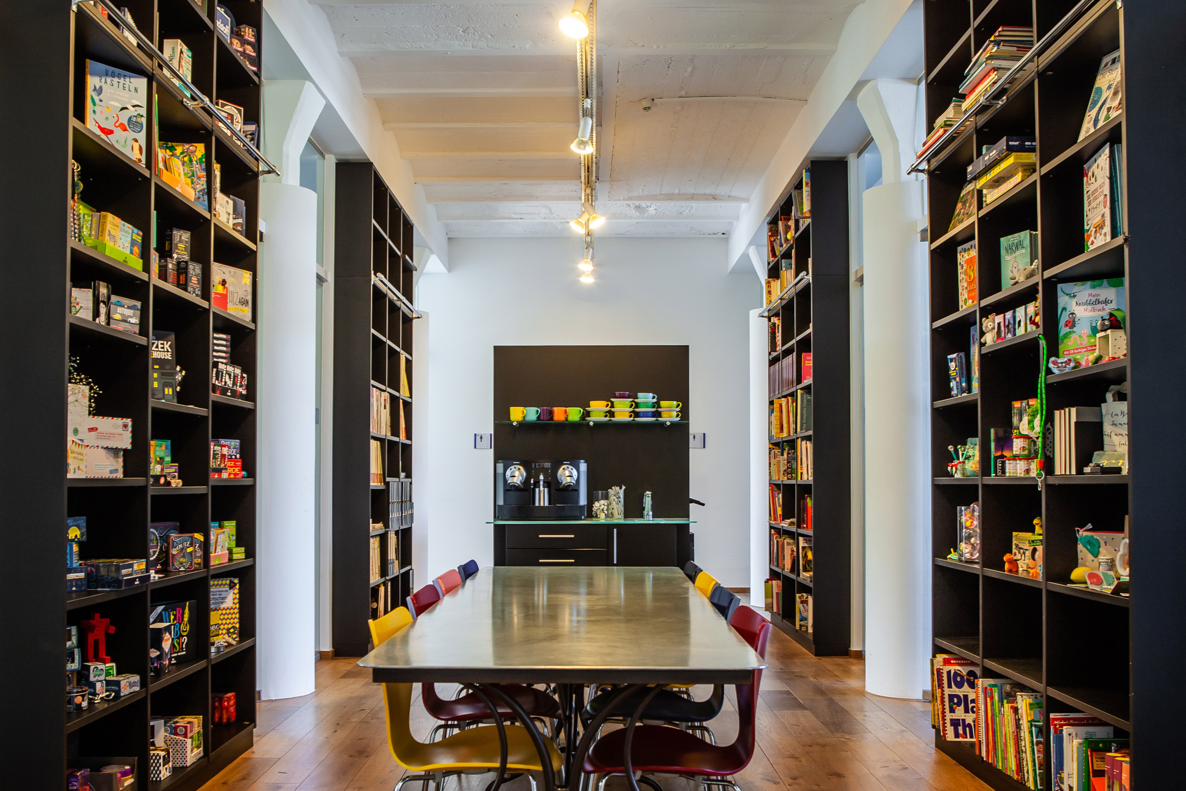 Verlagsflur mit langem Tisch, bunten Stühlen, Regalen voller Bücher und Kaffeemaschine, Marke: moses. Verlag