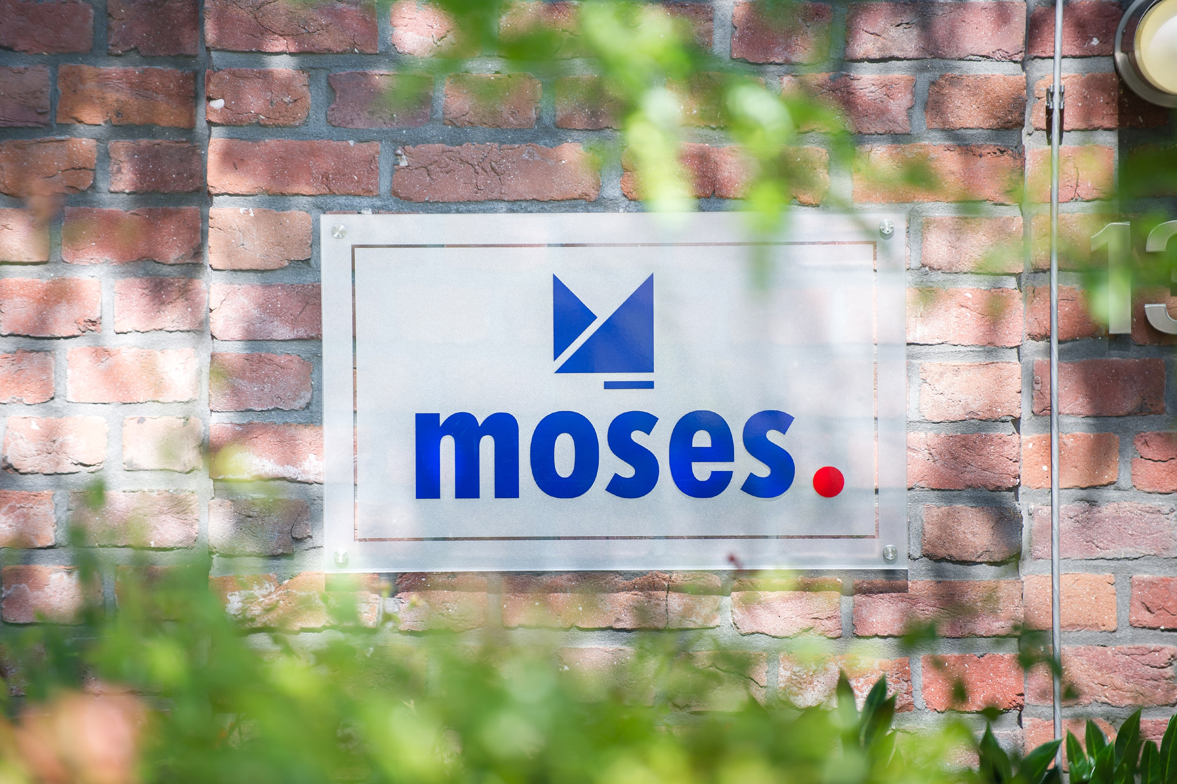 Firmenschild mit blauem moses. Logo und rotem Punkt auf Glas vor roter Backsteinwand Marke: moses. Verlag